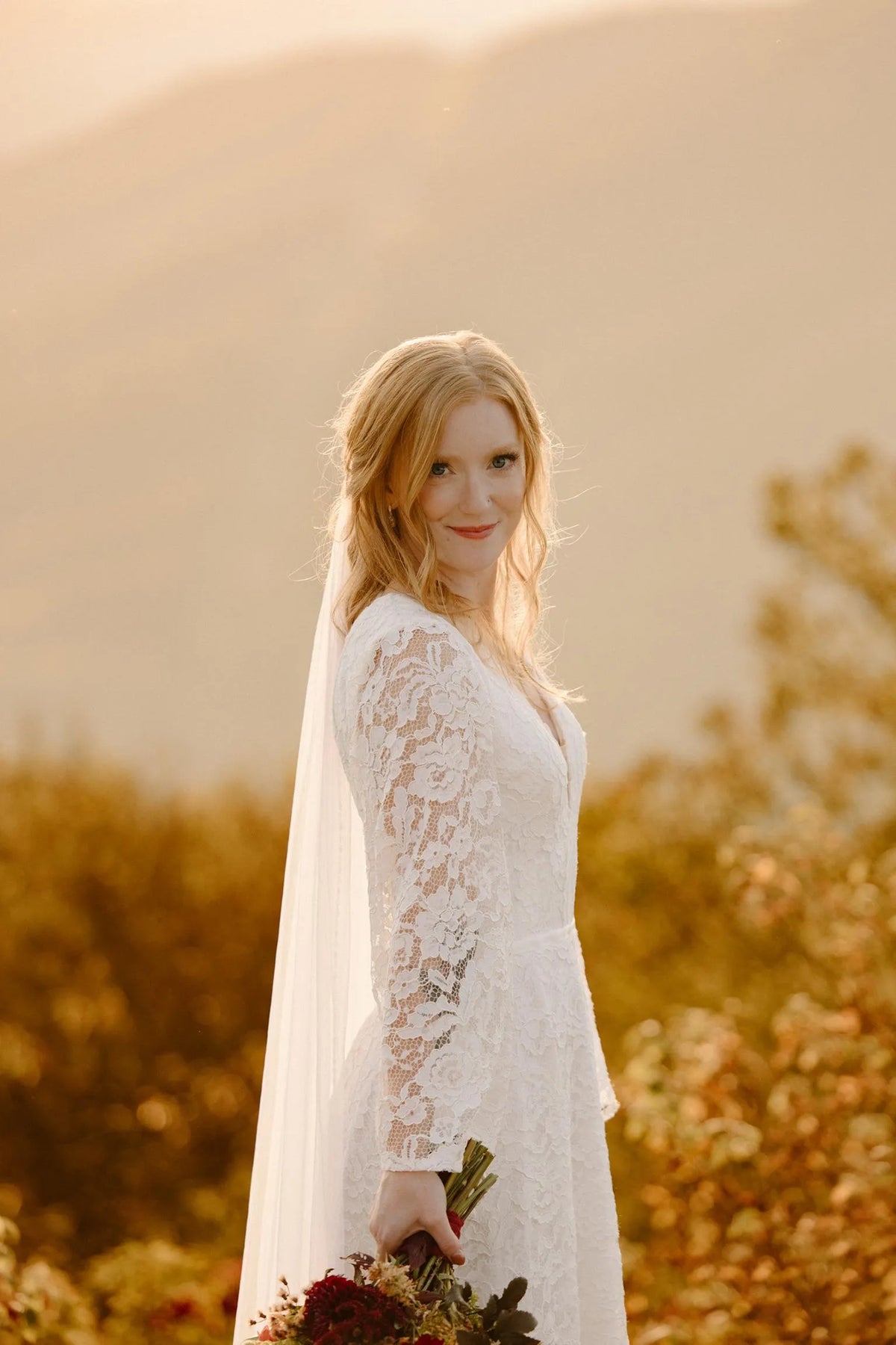 Bride outdoors in a long sleeve lace wedding dress with a veil and bouquet in warm evening light.