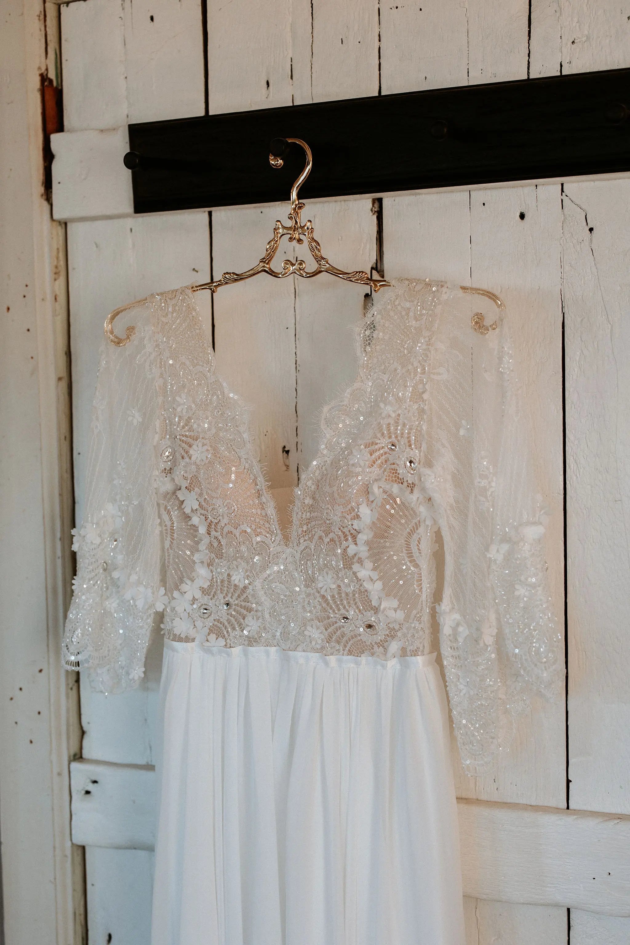 Boho lace wedding dress with long sheer embroidered sleeves, deep V neckline, floral lace bodice, and flowing chiffon skirt hanging on a rustic wooden door.